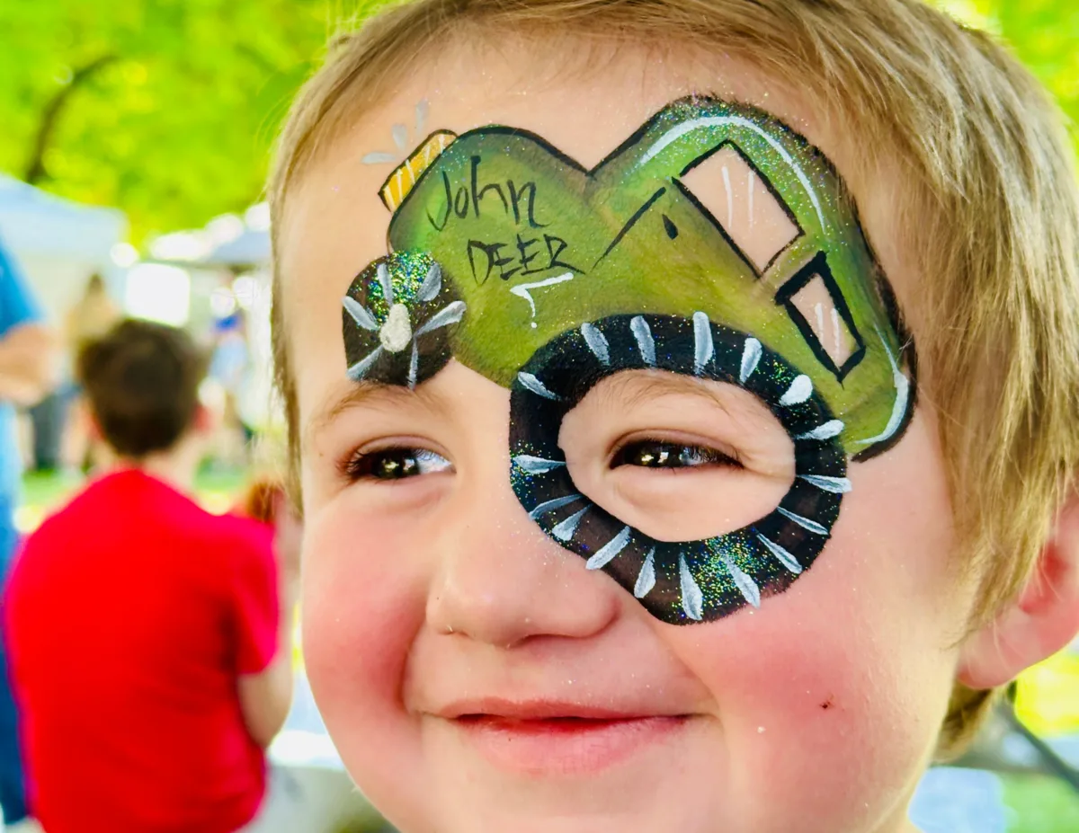 Child smiling with a green John Deere tractor face-painting design.
